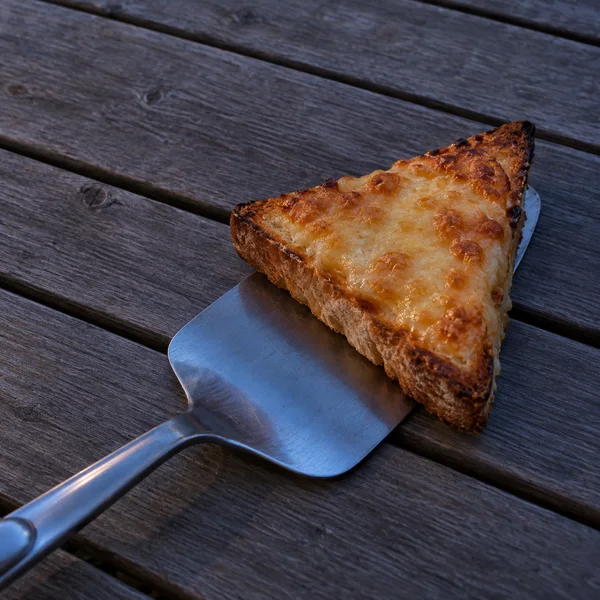 A triangular slice of browned cheese toast rests on a stainless-steel spatula on weathered wooden slats, with cool blue light reflected in the blade.