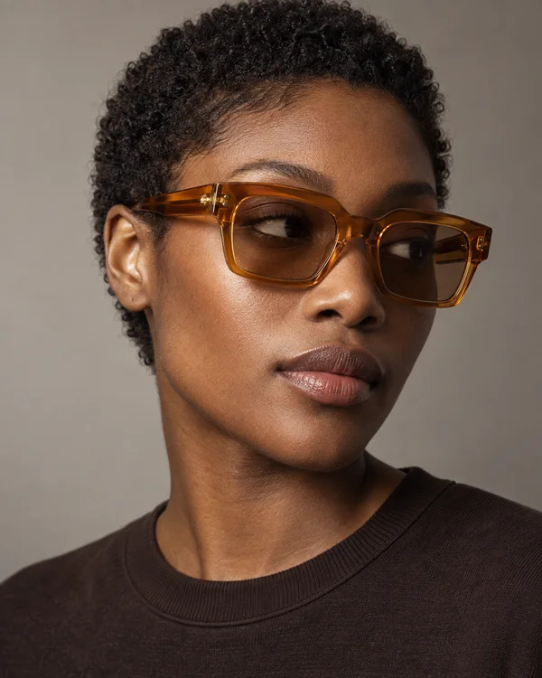 Close-up studio portrait of a dark-skinned model with short natural curls, wearing translucent amber rectangular sunglasses and a dark crewneck against a…