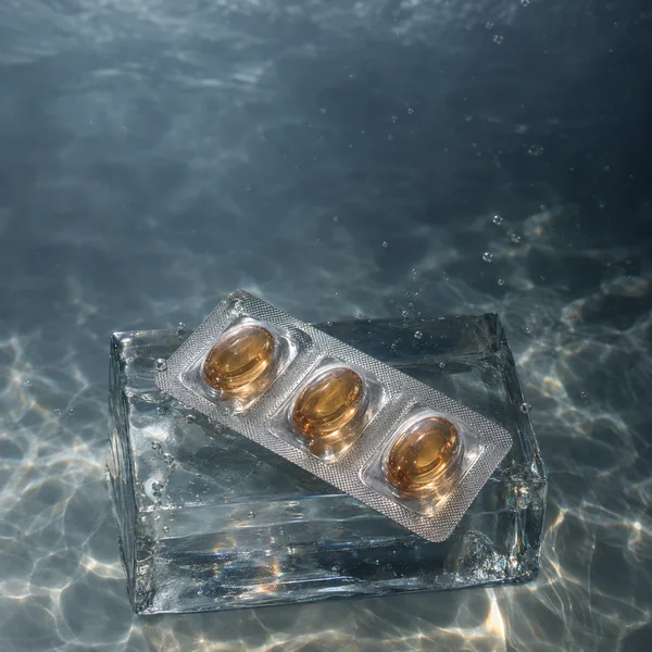 A diagonal silver blister strip holding three amber oval capsules rests on a clear glass block underwater, with bubbles and bright light patterns across the…