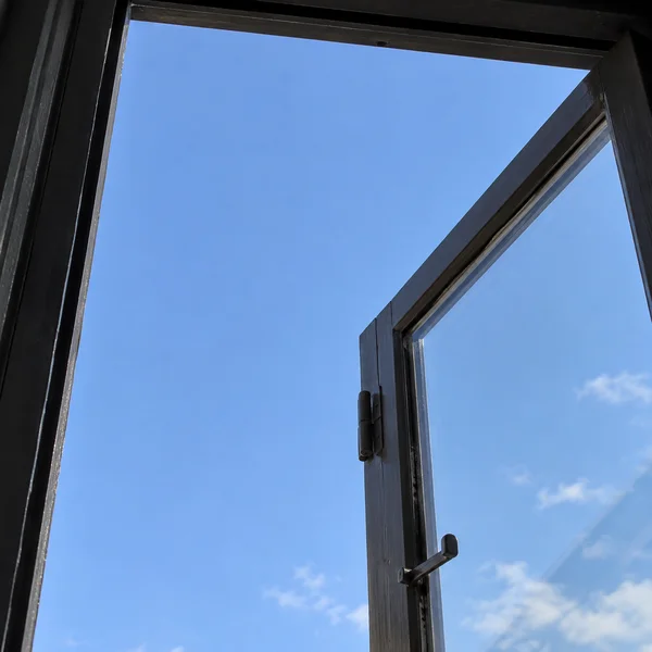 Upward view through an open dark-framed casement window, with its hinge and latch visible against a mostly clear blue sky.
