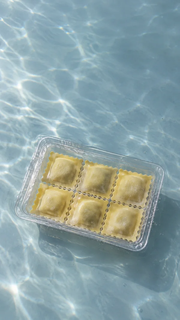 A clear rectangular tray holding six pale golden ravioli in two rows sits at an angle on a pale blue-grey underwater surface, with light patterns and small…