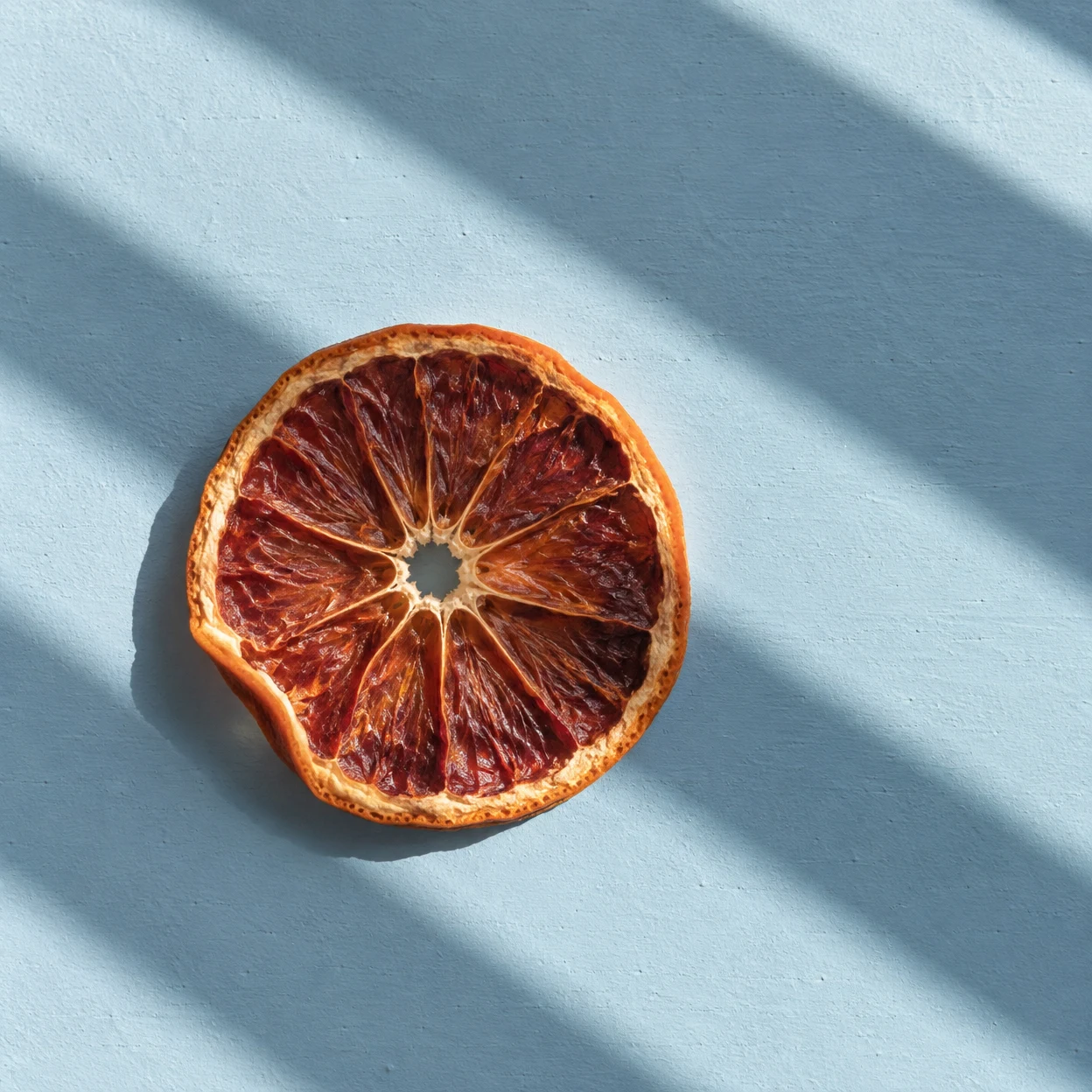 A dried blood-orange wheel lies on a pale blue painted tabletop, lit by diagonal bands of sunlight and shadow.