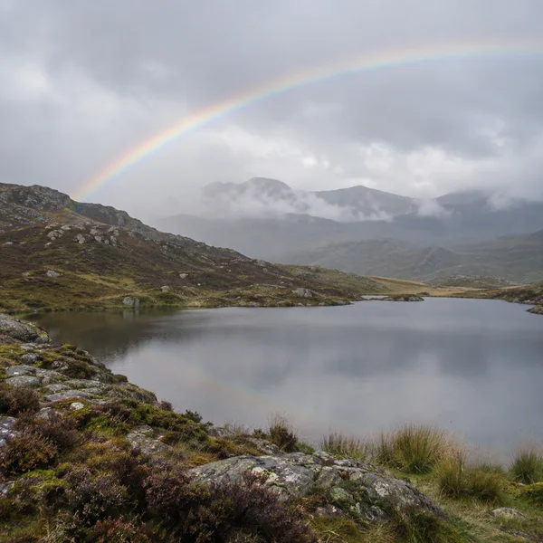 A soft rainbow arcs over a still upland tarn, with rocky heather-covered slopes in the foreground and mist-veiled ridges beneath a grey sky.