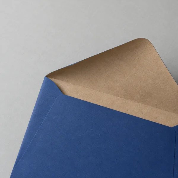 A dark blue paper mailer lies diagonally on a pale grey tabletop, opened to reveal a broad kraft-paper flap and empty interior; the mailer extends beyond the…