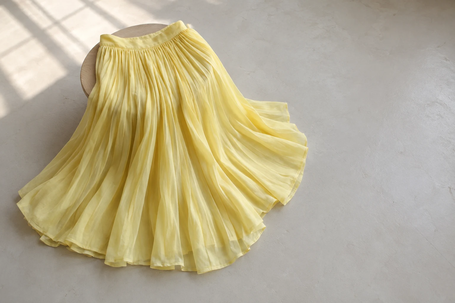 A lemon-yellow pleated skirt drapes over a mostly concealed round wooden stool and fans across a pale grey floor, with soft window-shaped light and open space…