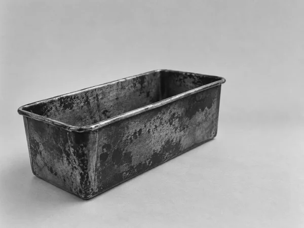 An empty, heavily mottled rectangular metal loaf tin sits diagonally on a plain light-gray surface, with its open interior and rolled rim visible against a…
