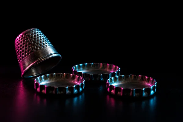 A silver sewing thimble tilted beside three unprinted metal bottle caps on a glossy black surface, edged with magenta and cyan light against a black background.