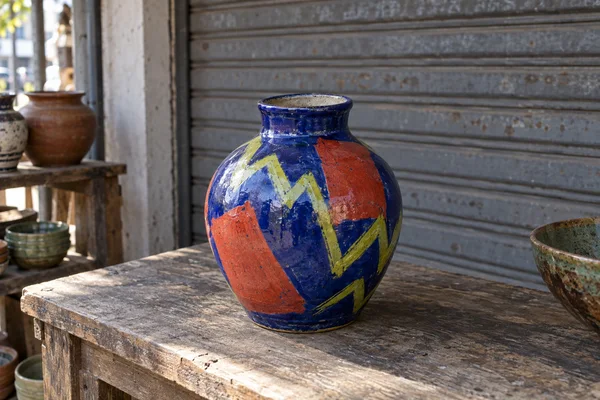 A large hand-painted ceramic vase with a cobalt-blue body, lime zigzags and vermilion blocks sits on a weathered wooden counter in front of a grey corrugated…