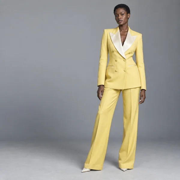 A full-length woman with close-cropped hair stands against a cool-grey studio backdrop in a pale yellow double-breasted suit with ivory satin lapels and…