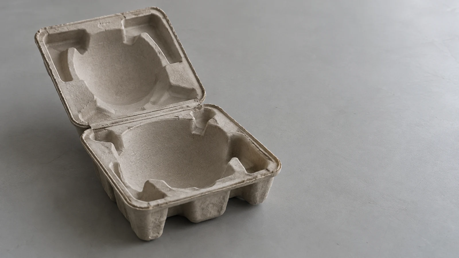 An open, empty warm-gray molded-pulp clamshell case with a rounded vessel-shaped cavity sits on a gray tabletop, viewed from above at an angle, with broad open…