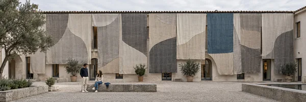 Ultra-wide courtyard view of a pale building draped with large overlapping fabric panels in cream, gray, and muted blue.