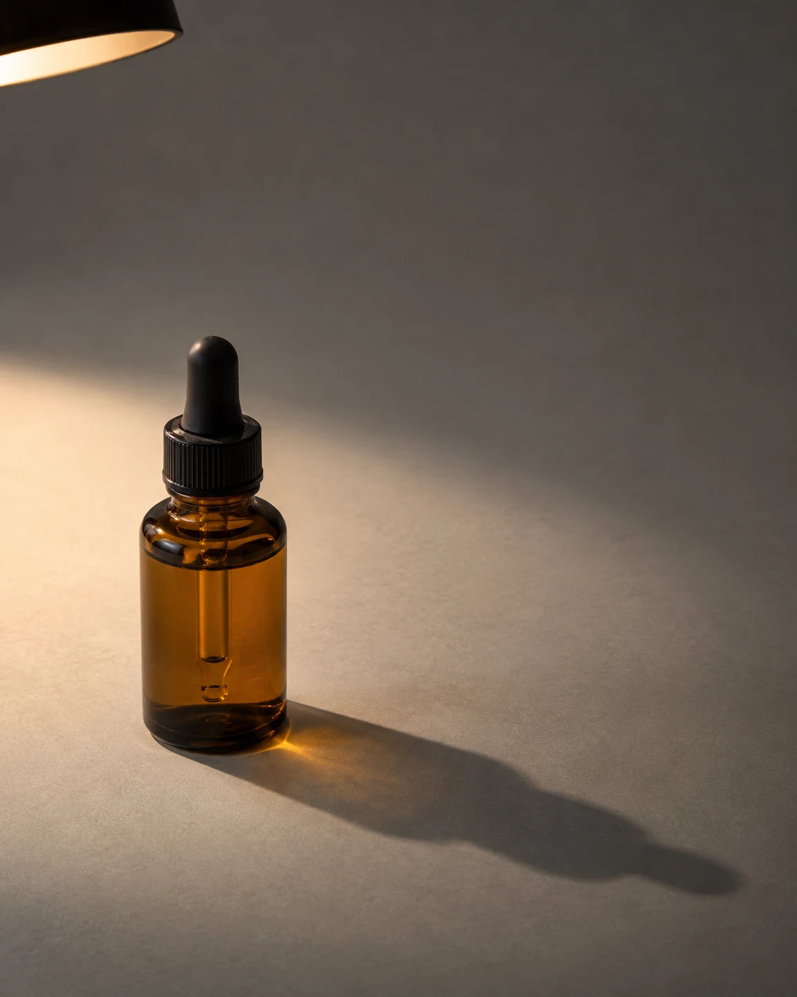 An unlabelled amber-glass dropper bottle stands on a cool-grey surface beneath the cropped edge of a black pendant shade.