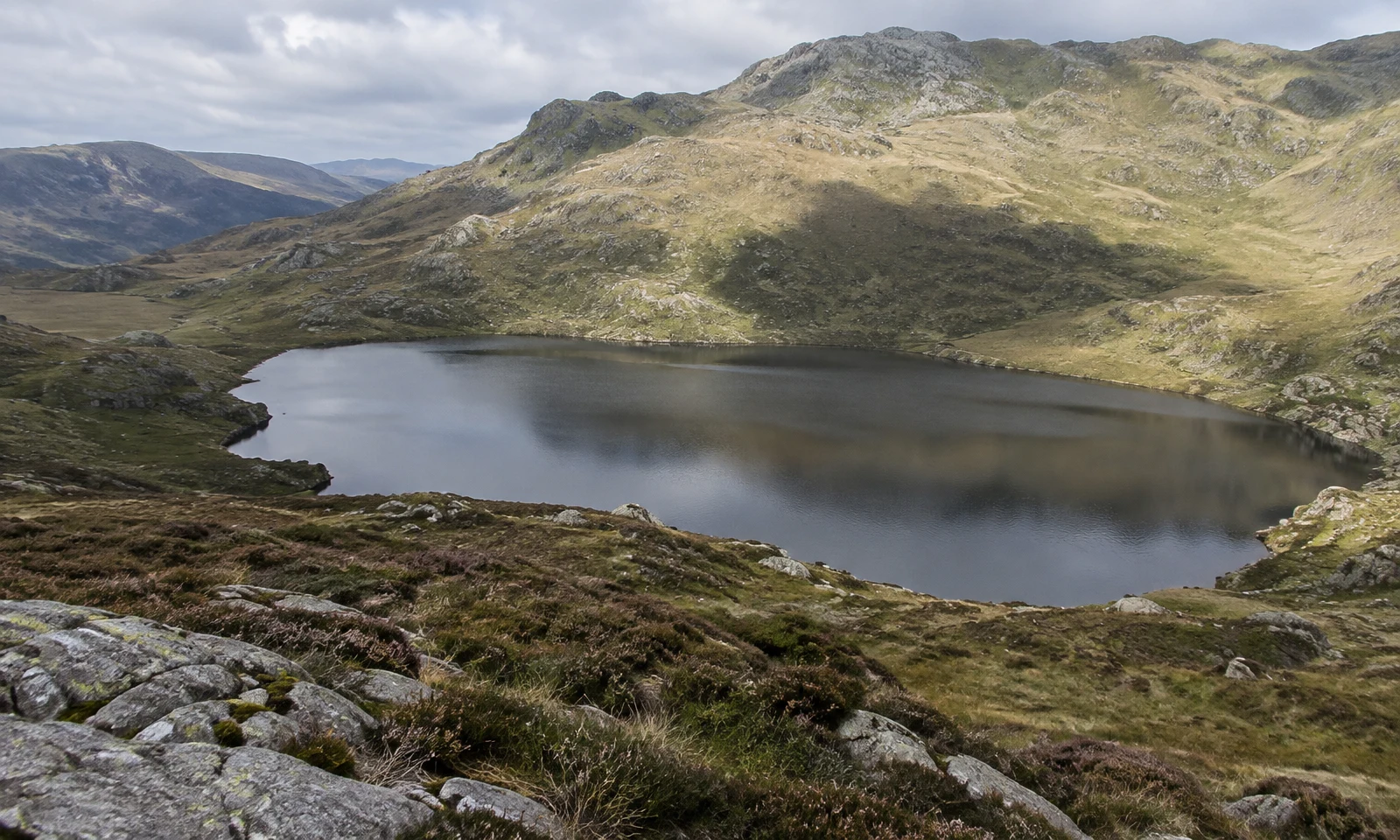 A still grey mountain tarn sits in a rocky upland basin ringed by heather and pale grass, with a broad shadow darkening the far slope and part of the water…