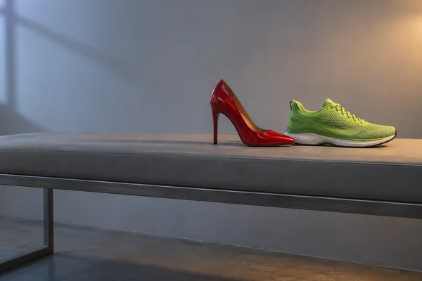 A glossy red high-heeled pump and a lime-green mesh running shoe sit on a long grey bench against a plain grey studio wall, with cool light from the left and a…