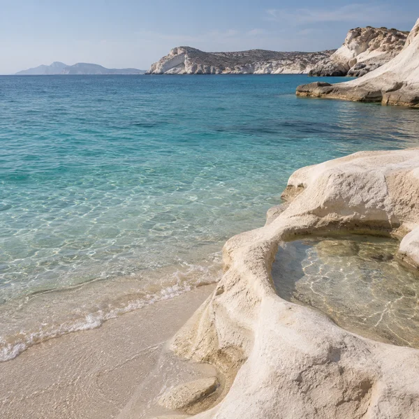 A pale sandy shoreline meets clear blue-green water beneath distant rocky island cliffs; a broad ivory rock ledge in the foreground contains a shallow pool…