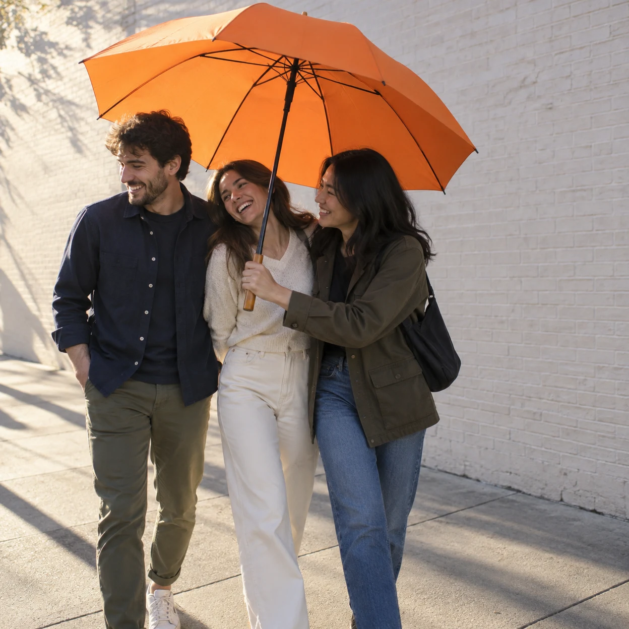 Three adult friends walk close together beneath a large orange umbrella beside a pale brick wall.