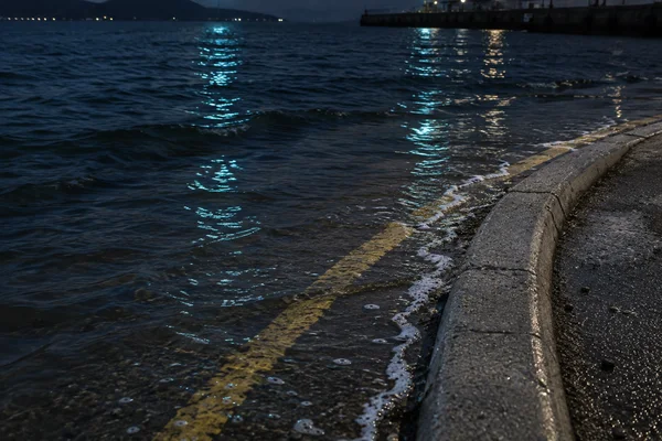 A curved concrete curb borders shallow water beside a yellow road-edge stripe, with a narrow foam line and cyan and warm reflections across the dark ripples…