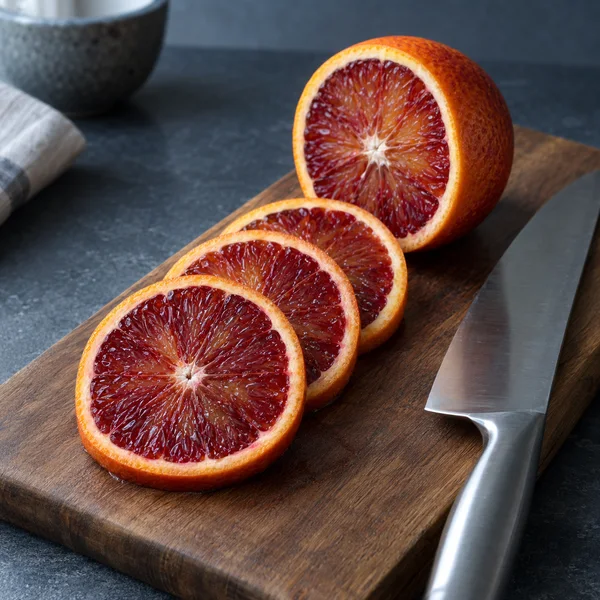 Three overlapping blood-orange slices sit on a walnut cutting board in front of a cut half-orange, with a stainless-steel knife alongside them.