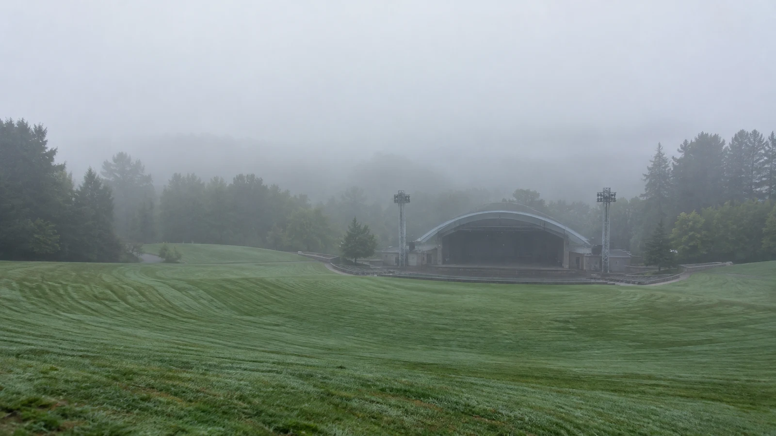 An empty, gently sloping lawn with curved mowing stripes leads toward a low arched concert stage and two floodlight towers, with trees fading into dense…