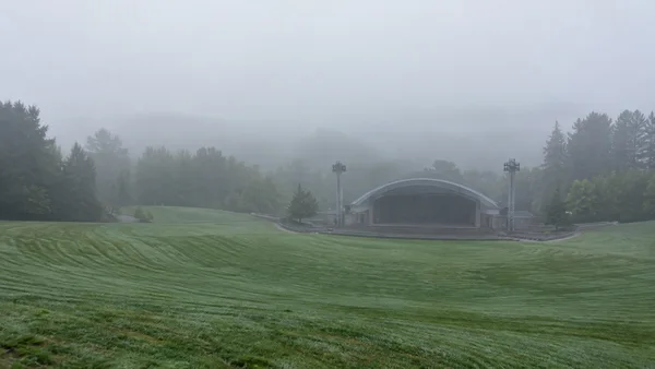 An empty, gently sloping lawn with curved mowing stripes leads toward a low arched concert stage and two floodlight towers, with trees fading into dense…