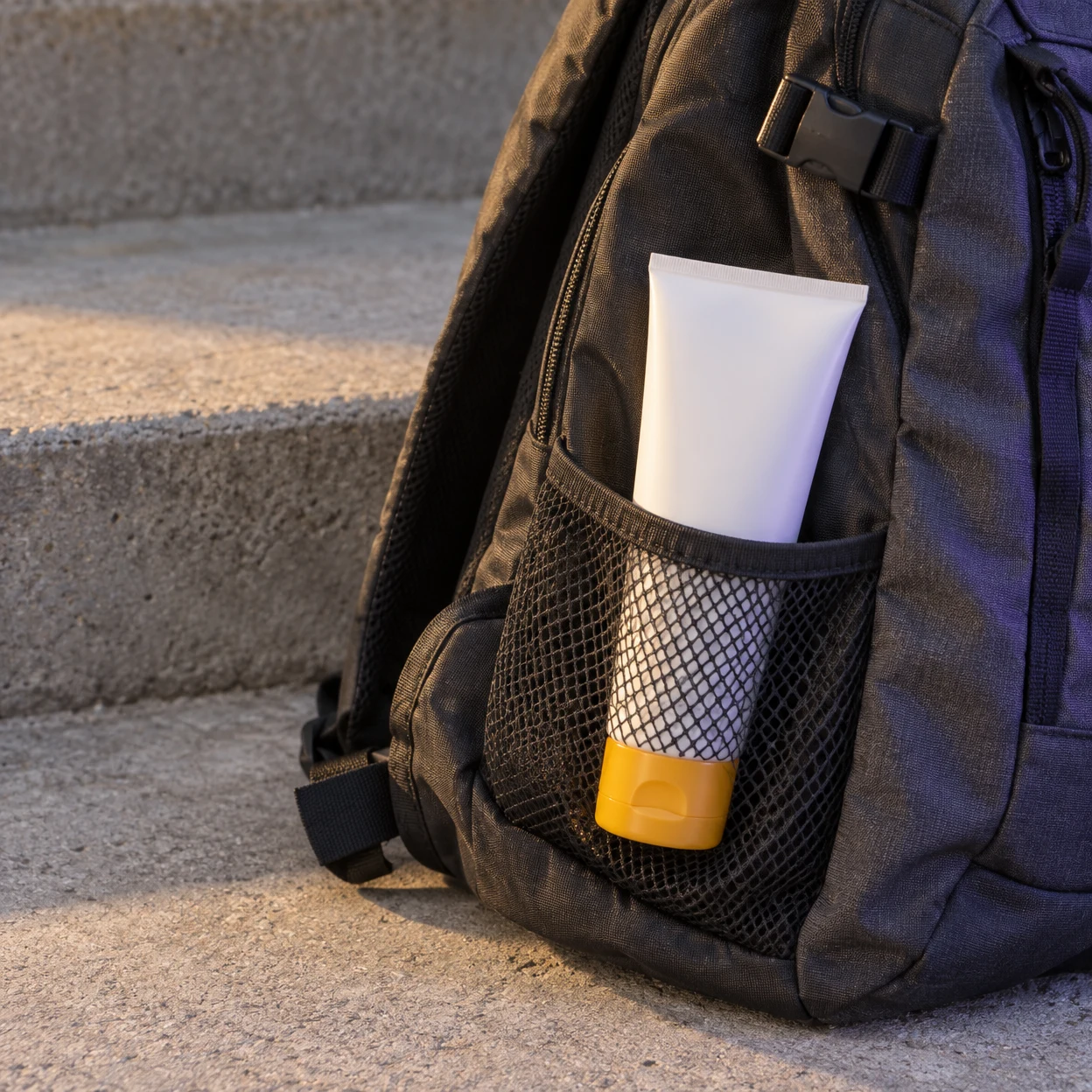 A charcoal daypack rests on rough concrete steps, with a plain white squeeze tube and yellow cap pointing downward in its mesh side pocket.