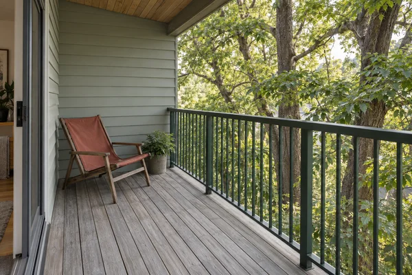 A rust-red canvas sling chair sits on a weathered timber balcony beside pale sage siding and a dark-green metal railing, with leafy trees and thick trunks…