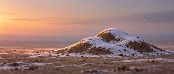 A low, irregular brown earth mound with patchy white snow sits on a rocky plain at sunset, with sparse snowflakes in the air and open sky to the left.