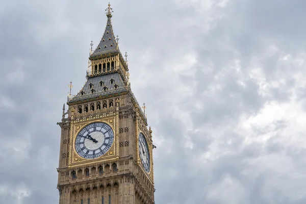 A close upward view of Elizabeth Tower shows two clock faces, ornate stonework, gilded details and its dark pointed roof against a cloudy sky.
