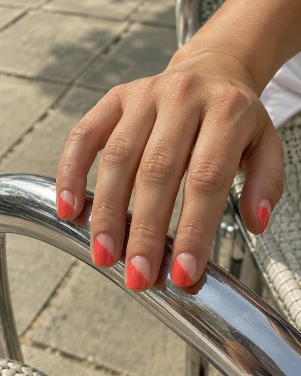 A hand with coral-red diagonal nail art rests across a curved chrome café-chair arm, with pale stone paving and part of a woven chair seat in the background.