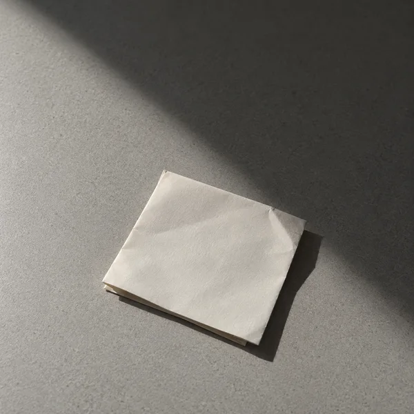 A blank off-white folded sheet rests below centre on a grey textured tabletop, lit by a hard diagonal beam and casting a crisp shadow to the lower right.