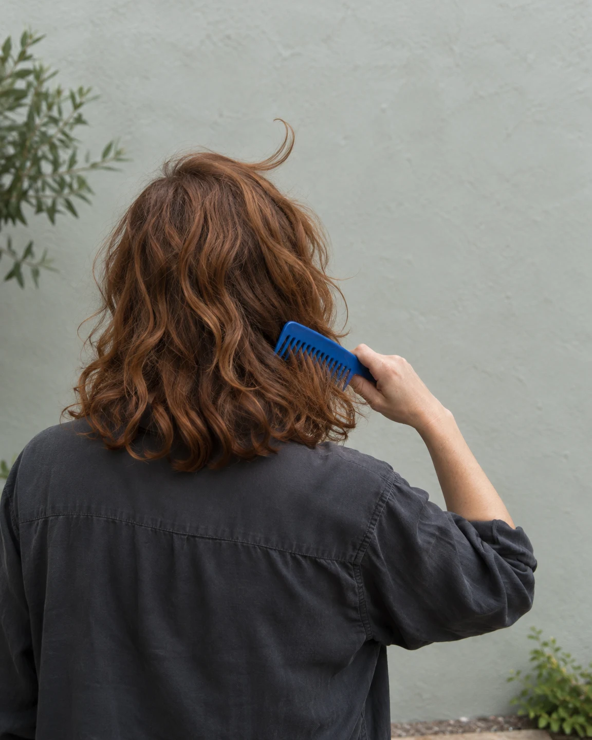 Rear view of an adult with shoulder-length copper-brown wavy hair, drawing a cobalt-blue wide-tooth comb through the hair while standing before a pale…