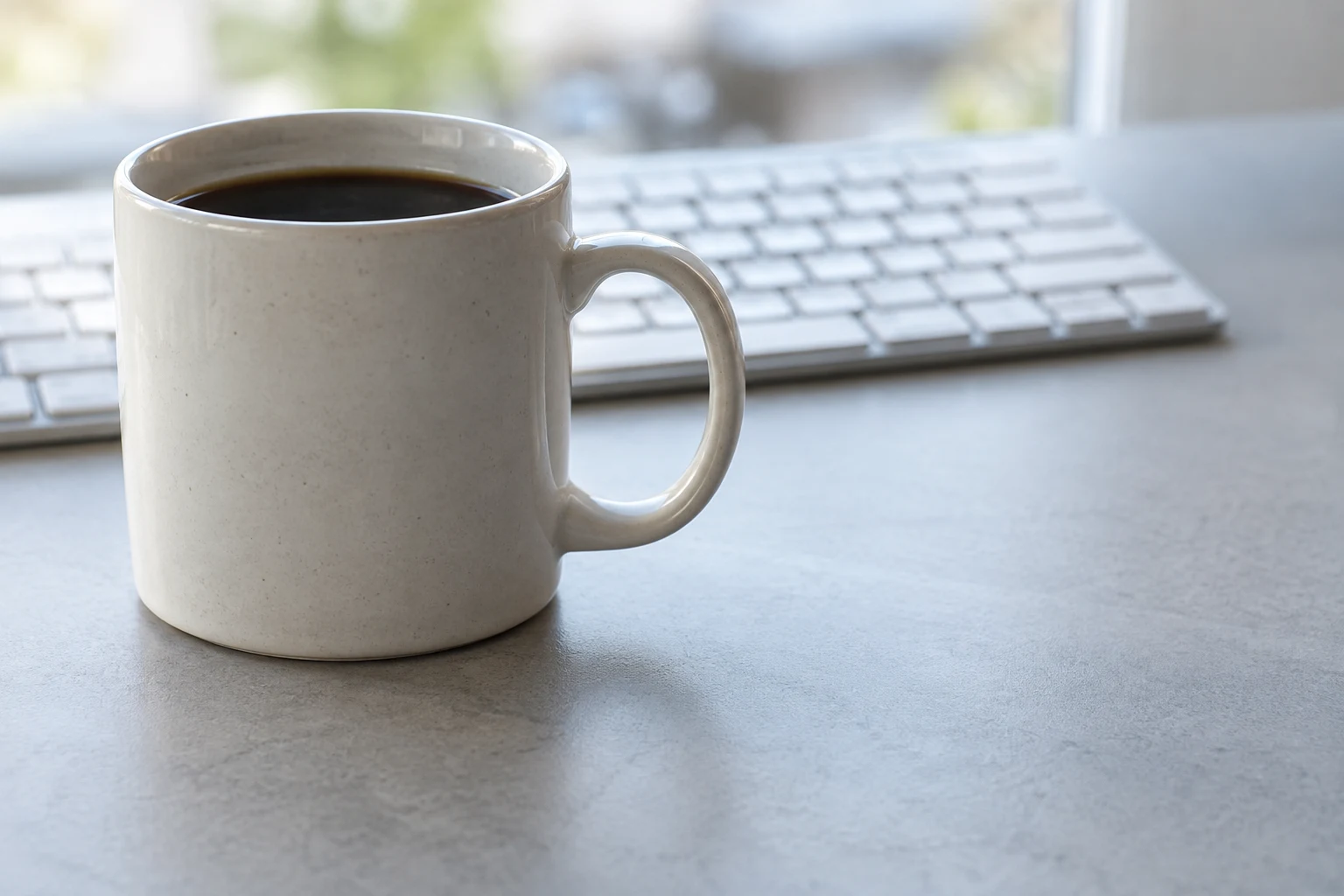 An off-white ceramic mug filled with dark coffee sits left of centre on a pale grey desk, its right-facing handle in front of a softly blurred keyboard.