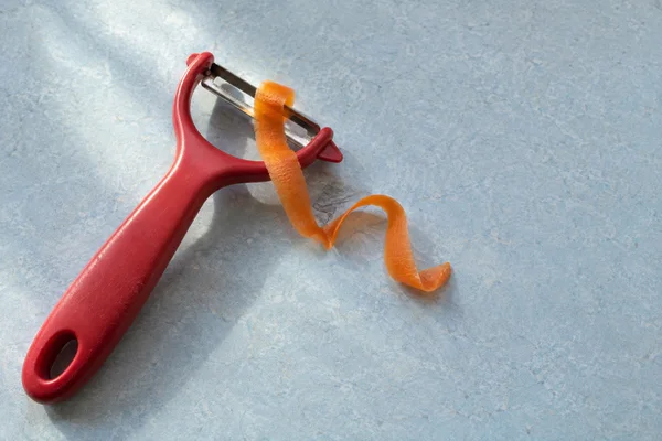A red-handled stainless-steel Y-peeler lies diagonally on a pale blue mottled counter, with an orange carrot ribbon caught at the blade and curling beside it.