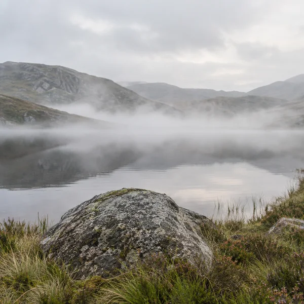 A large speckled granite boulder sits among tussock grass and heather beside a still upland tarn, with low mist drifting across the water and hazy hills…