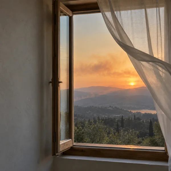 An open timber casement window and sheer curtain frame layered hills beneath an orange sunset, with trees and a strip of water visible in the distance.