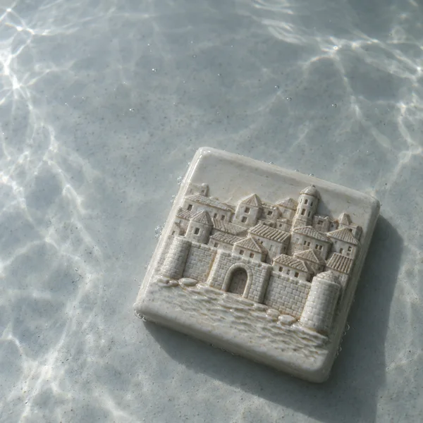 An ivory glazed ceramic tile with raised relief of a walled city, an arched gateway, towers and clustered roofs, rests diagonally on a pale gray tank floor…