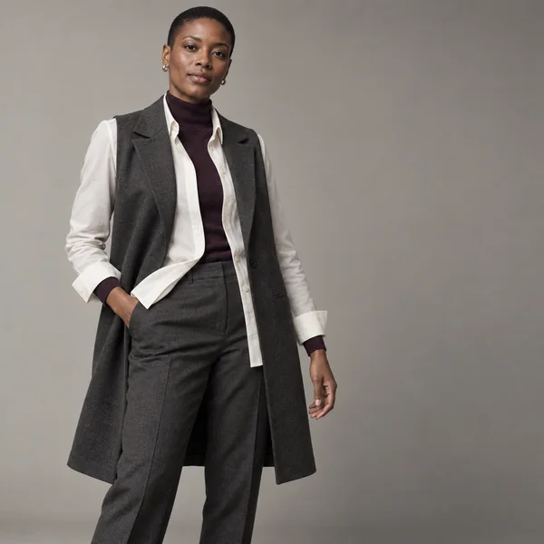 A short-haired woman poses against a warm-gray studio backdrop in a plum turtleneck, ivory shirt, charcoal sleeveless longline blazer and matching trousers…