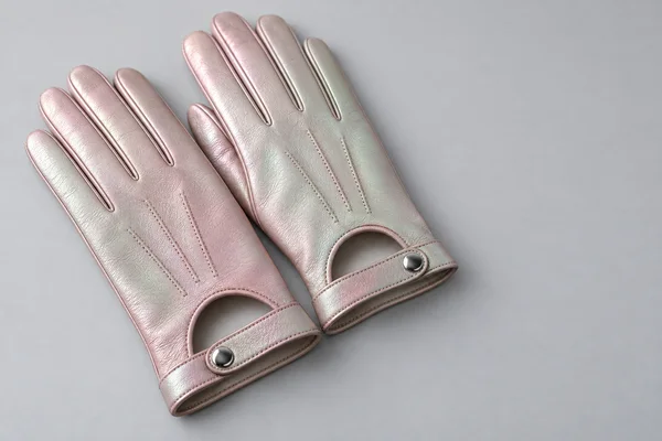 Two pearlescent leather gloves with finger seams, arched wrist openings, straps, and round metal snaps rest on a pale-gray surface, leaving open space to the…