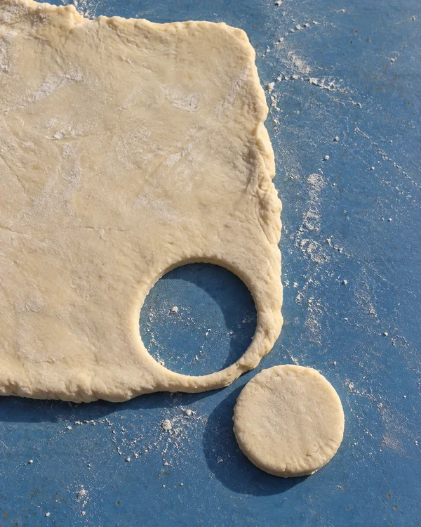 Overhead view of a pale sheet of biscuit dough with a circular hole and a separate round of dough beside it on a blue, flour-dusted tabletop.