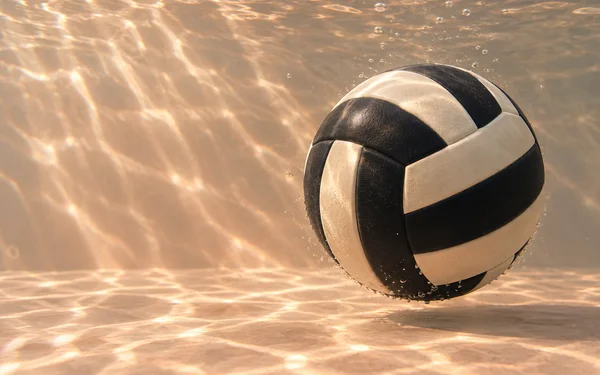 A black-and-ivory paneled rubber ball floats above a peach-colored floor in clear, warmly lit water, with small bubbles around it and broad bands of light…