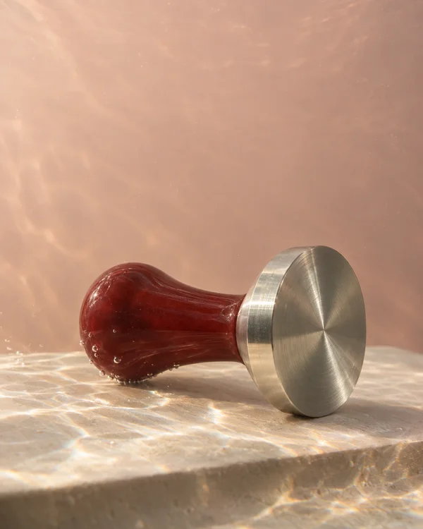 A stainless-steel espresso tamper with a dark red resin handle lies on its side on a pale textured ledge, surrounded by clear water against a blush-pink…