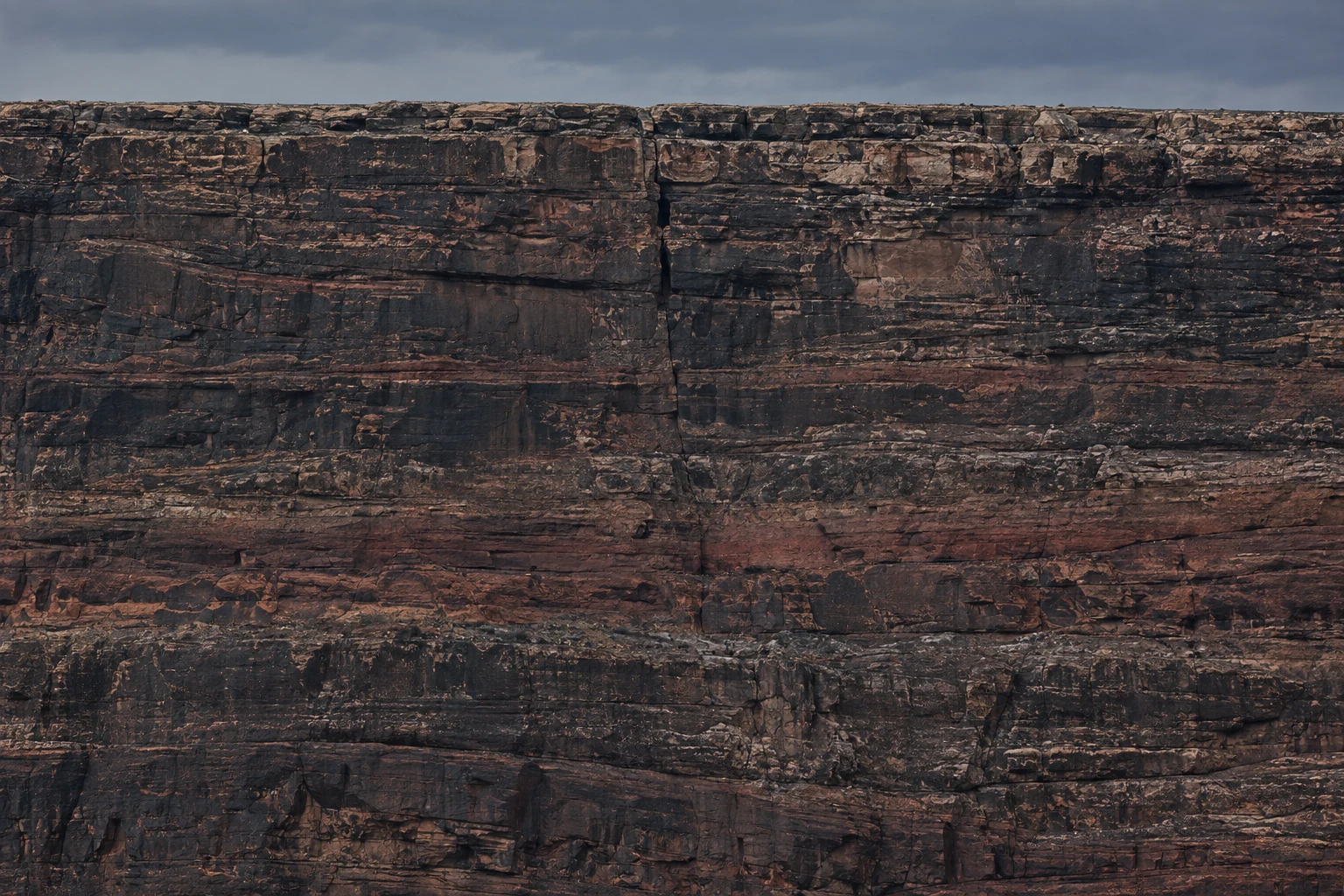 A close, face-on view of a broad mesa cliff beneath a narrow band of gray-blue sky, with dark horizontal rock layers and a prominent vertical fissure near the…