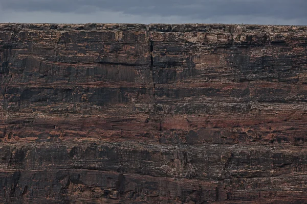 A close, face-on view of a broad mesa cliff beneath a narrow band of gray-blue sky, with dark horizontal rock layers and a prominent vertical fissure near the…