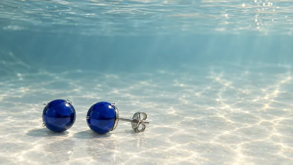 Two cobalt-blue spherical stud earrings rest on a pale submerged floor, with one facing the camera and the other lying sideways to show its metal post and…