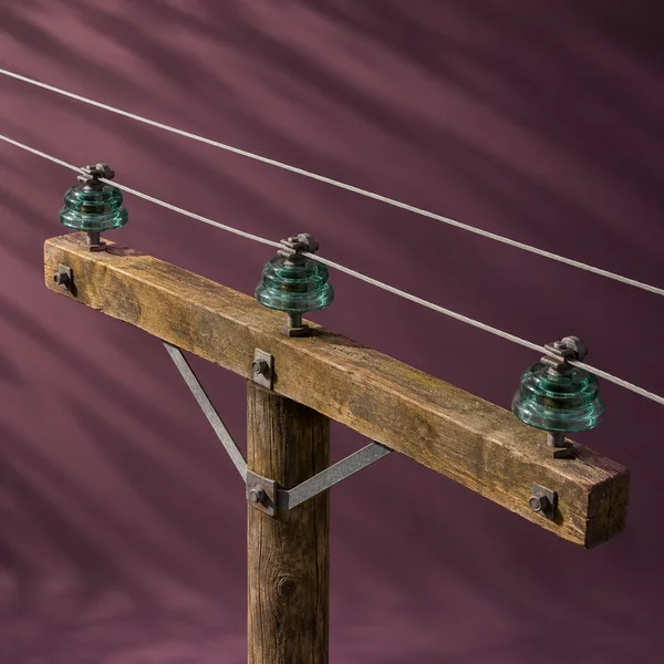 A weathered wooden utility crossarm on a short pole section carries three green glass insulators and metal fittings against a plum backdrop crossed by broad…
