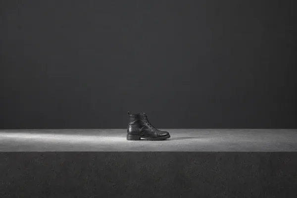 A single black leather lace-up ankle boot stands near the center of a broad gray platform against a charcoal wall, lit by a bright sweep of light across the…