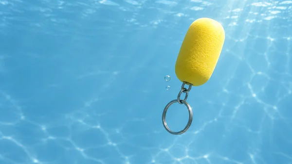 A lemon-yellow foam capsule floats underwater, connected by several metal loops to a split keyring hanging below it; two small bubbles sit beside the connector…