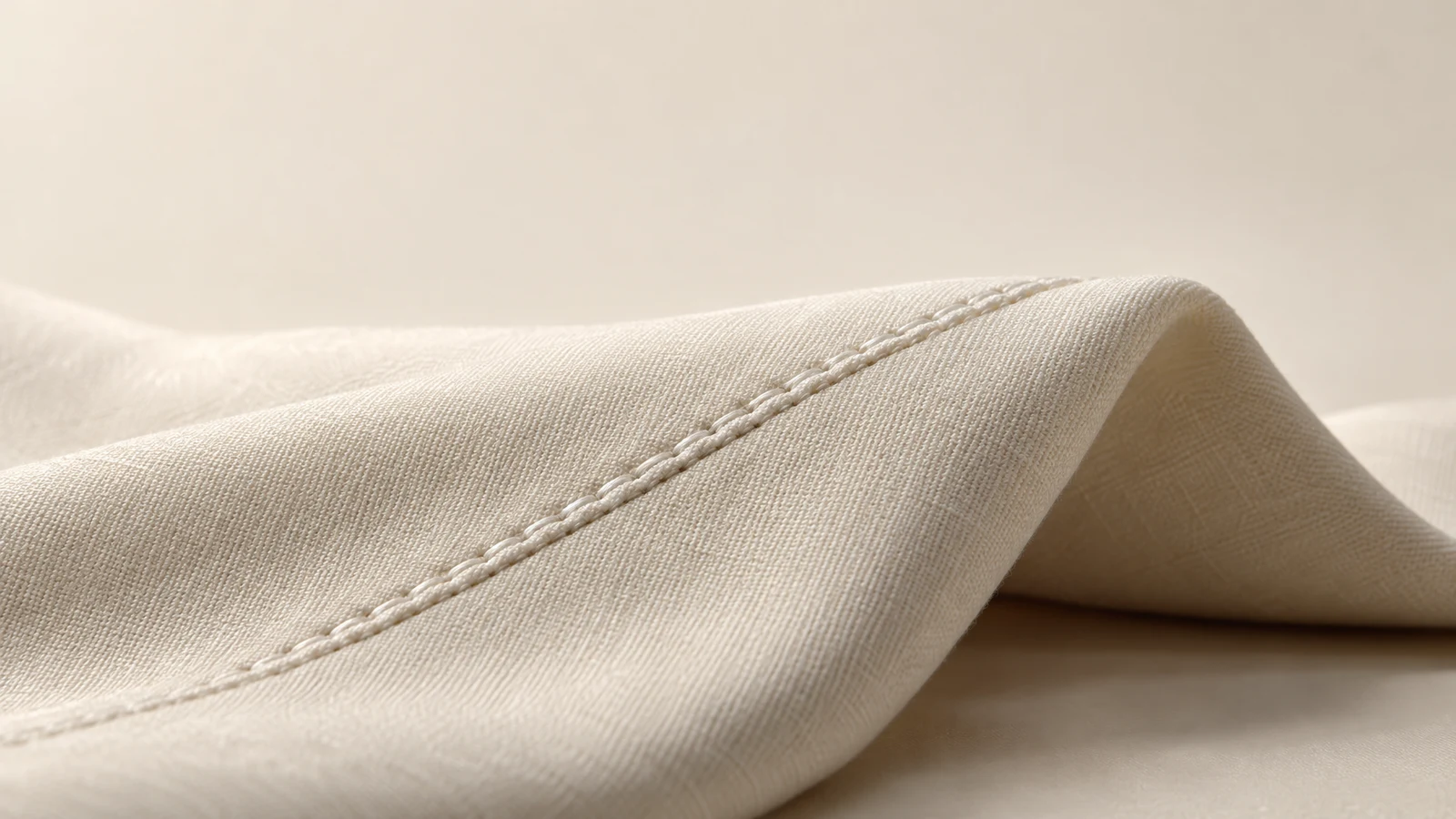 A close-up of ivory woven fabric folded into a broad raised ridge, with a diagonal seam crossing the foreground and warm soft shadows beneath the fold.