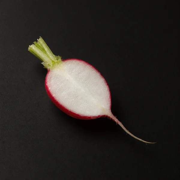 A lengthwise half of a red radish rests cut-face up on a dark charcoal surface, with its green stalk angled toward the upper left and slender root extending…