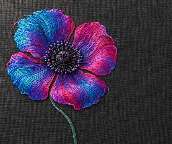 A large four-petaled poppy embroidered in cyan, violet, and magenta thread sits left of center on charcoal linen, with a curved stem and open dark fabric to…